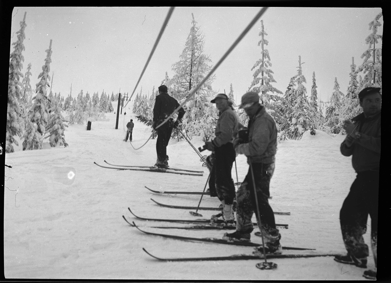 A handful of unidentified people stand in a line for the ski lift at Lookout Pass. They are all wearing ski gear and the ground and trees are covered in snow.