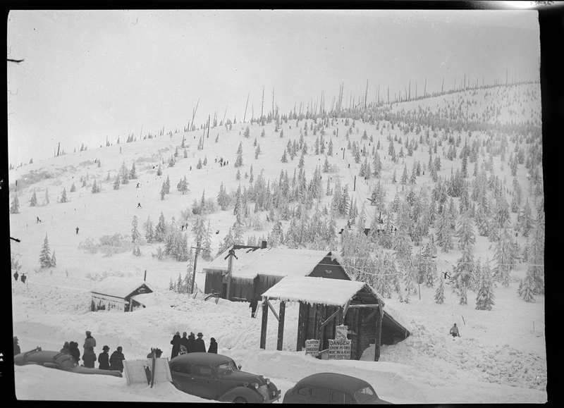 Photo of a group of people standing behind parked cars in the snow at Lookout Pass. There are a few buildings behind them, and snow covered trees are visible behind them.
