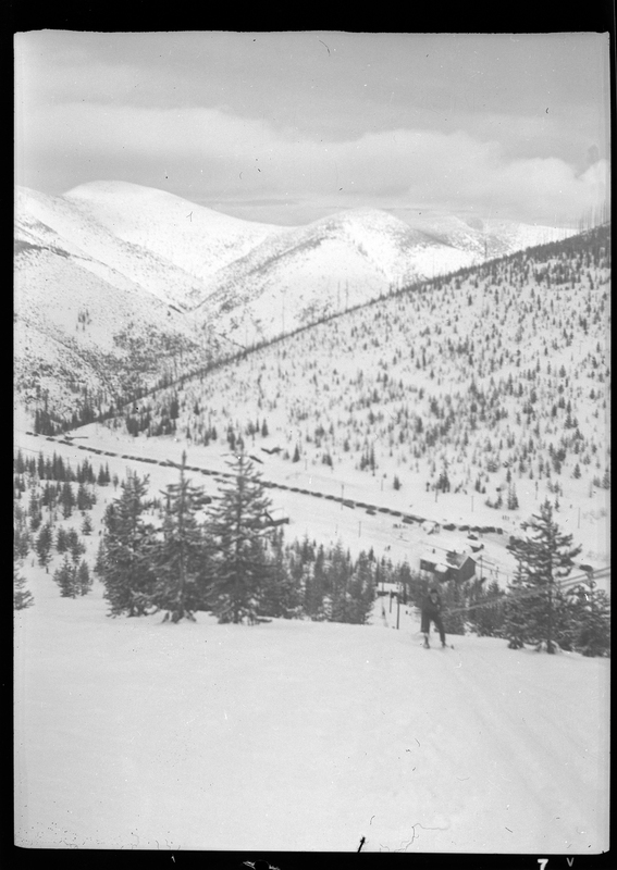 An unidentified person is using the ski lift at Lookout Pass. The ground is covered in snow and looking down the hill trees and a line of parked cars are visible.