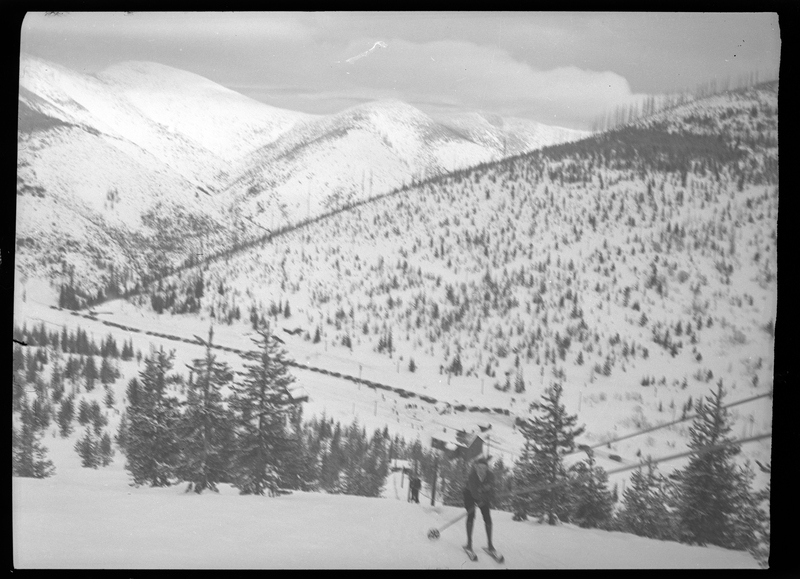 An unidentified person is using the ski lift at Lookout Pass. The ground is covered in snow and looking down the hill trees and a line of parked cars are visible.