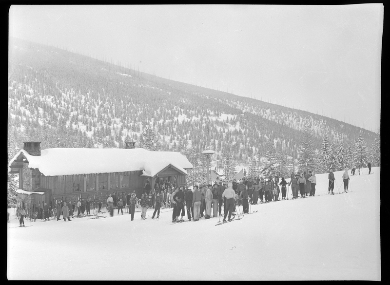 A large group of people in ski gear are gathered together outside for a skiing tournament. The ground and the building that everyone is gathered around is covered in snow, and trees can be seen in the background.