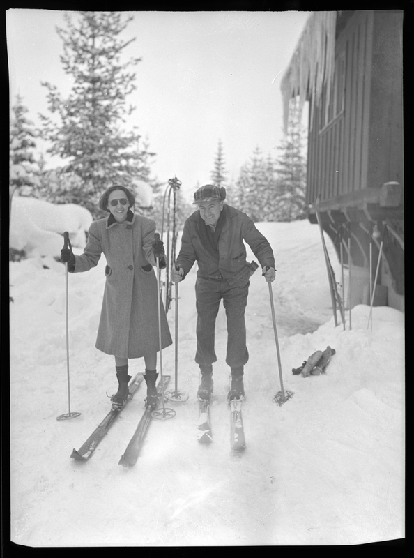 An unidentified man and woman wearing ski gear stand next to a building in the snow for a tournament. They are both smiling at the camera.