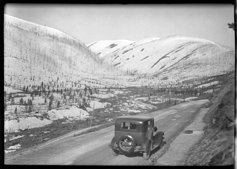 Photo of a car driving away from the photographer down a snow and mud covered road at Lookout Pass. There is a wall of snow piled up on one side of the road and trees can be seen in the background.