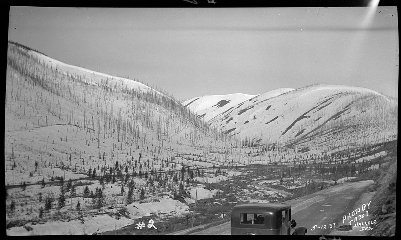 Photo of a car driving away from the photographer down a snow and mud covered road at Lookout Pass. There is a wall of snow piled up on one side of the road and trees can be seen in the background. Only the top of the car is visible.