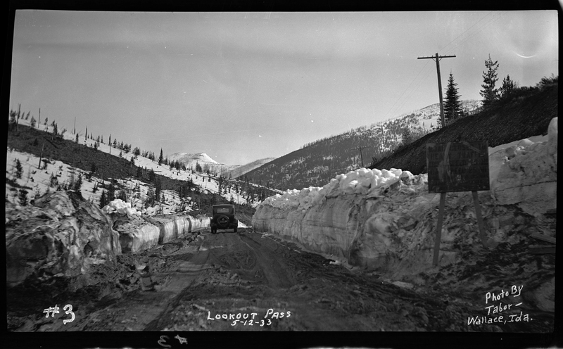 Photo of a car driving away from the photographer down a snow and mud covered road at Lookout Pass. There are walls of snow piled up on either side of the road, a sign propped up in the snow, and trees can be seen in the background.