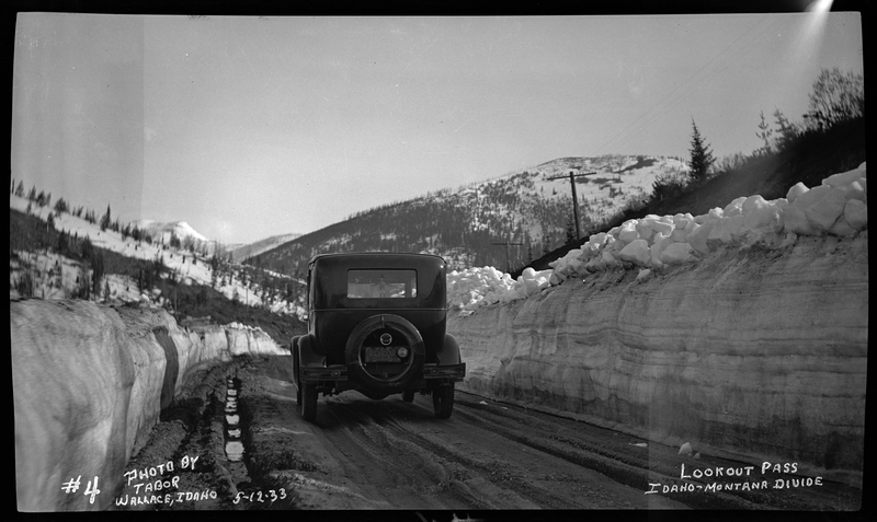 Photo of a car driving away from the photographer down a snow and mud covered road at Lookout Pass. There are walls of snow piled up on either side of the road and trees can be seen in the background.