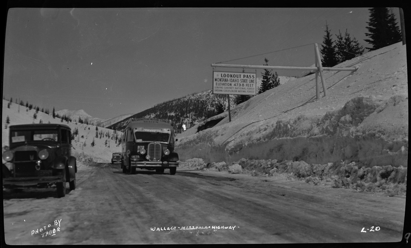 Two cars are driving down the road, towards the photographer, on the Wallace-Missoula Highway. There is a car parked on the side of the road. There is also a sign for Lookout Pass on the other side of the road that reads, "Lookout Pass; Montana-Idaho State Line; Elevation 4738 Feet; Leaving Cabinet National Forest; Entering Coeur d'Alene National Forest." 