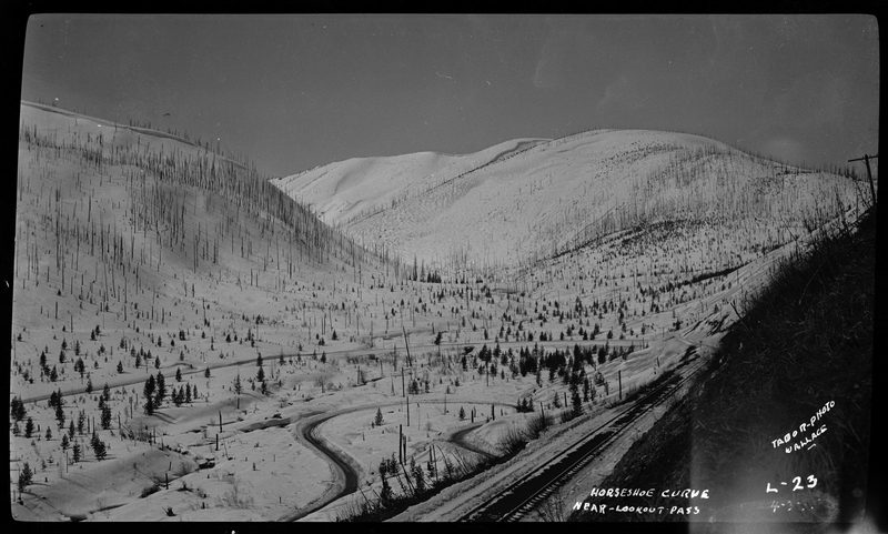 Photo of Horseshoe Curve near Lookout Pass in the snow. Horseshoe Curve appears to be a road in the landscape that curves dramatically. There are trees, hills, and a railroad track all visible in the photo.