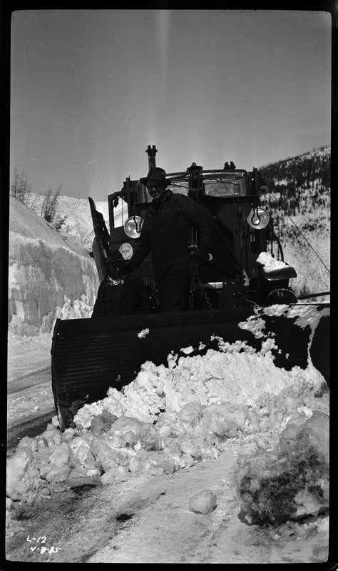 An unidentified man is seen standing on the front of a snow plow at Lookout Pass. There is snow on the ground in front of the snowplow, and in the background.
