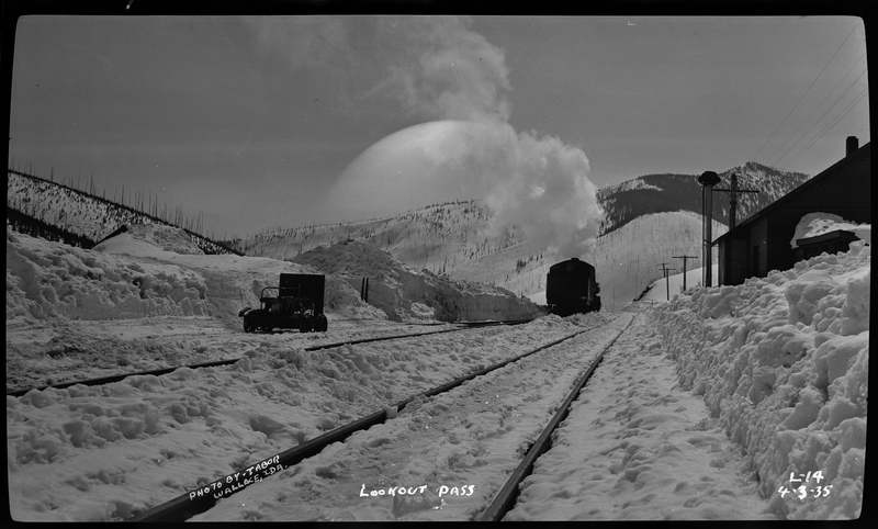Photo of snow covered railroad tracks at Lookout Pass. There is a train engine on one of the tracks, and a building off to the side.
