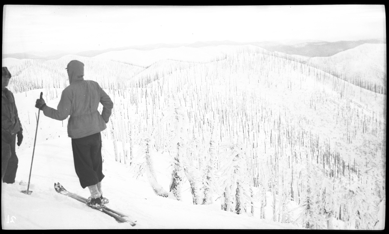 Photo of two skiers at Lookout Pass talking to each other. One is wearing skis and holding poles, while it is unclear if the other unidentified person has either. In the background, a snow covered hill with trees scattered on it is visible.