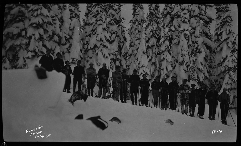 Photo of a group of unidentified people standing in a line for a photo. They are all wearing ski gear. The ground and trees around them are covered in a heavy layer of snow.