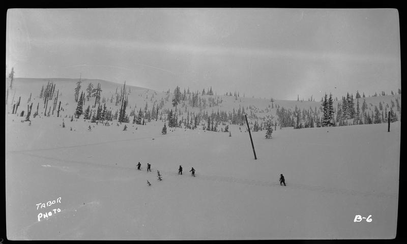 A group of unidentified people ski in a line down the hill at Lookout Pass. The ground is covered in snow and trees are in the background.