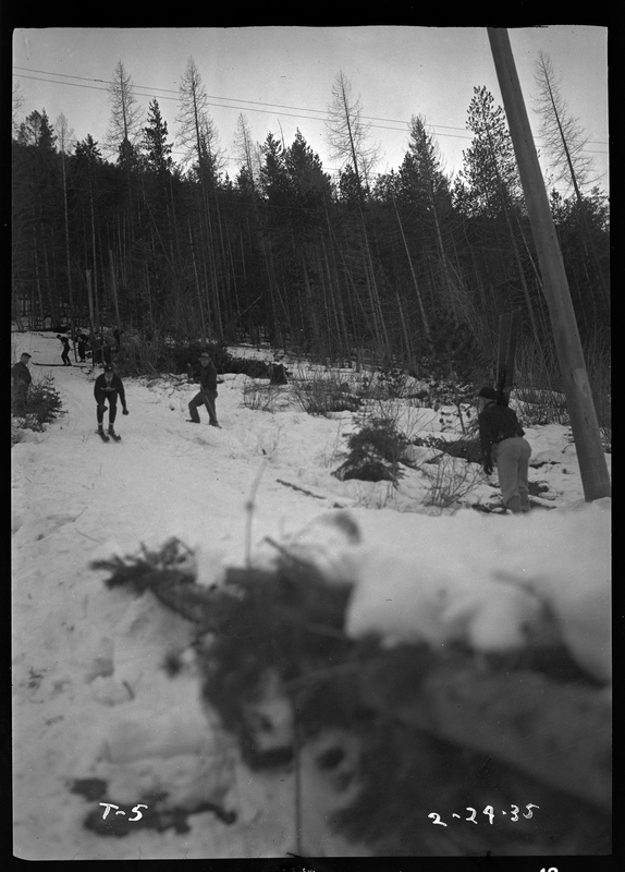 An unidentified person is seen skiing down the hill for the ski jump event at Lookout Pass. There are other people off to the side, and trees in the background.