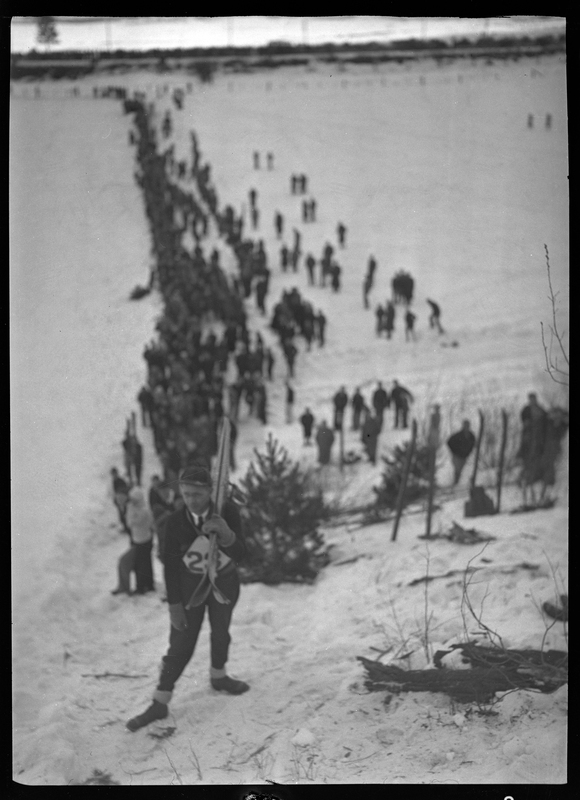 A line of people seemingly competing in the ski jump at Lookout Pass. Only one man is in focus in front of the photographer, and he is holding skis.