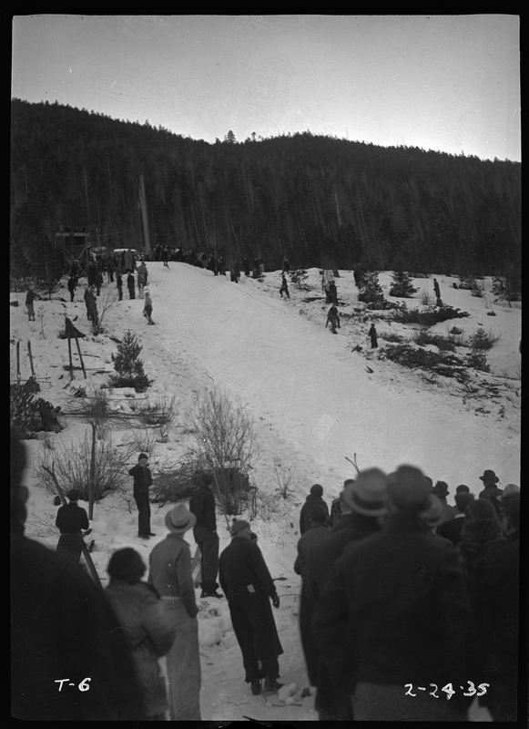 Photo of the ski jump hill located at Lookout Pass for a ski jump event. There is a ground of people at the base and a few people on the sides of the hill.