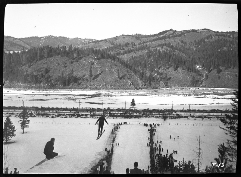 An unidentified person is mid jump at a ski jump event at Lookout Pass. There is snow on the ground and a crowd of people watching.