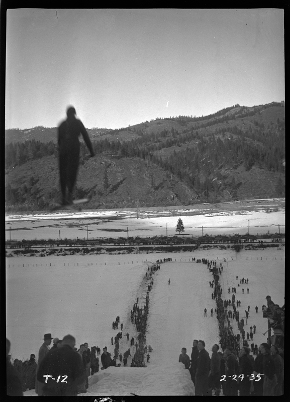 An unidentified person is mid jump at a ski jump event at Lookout Pass. There is snow on the ground and a crowd of people watching.