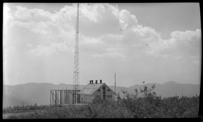 Photo of the radio beam station building at Mullan Pass. The building is in the distance, and a car can be seen parked next to it. There is a tall radio tower next to the building. The roof of the building has "Mullan Pass" written on it.