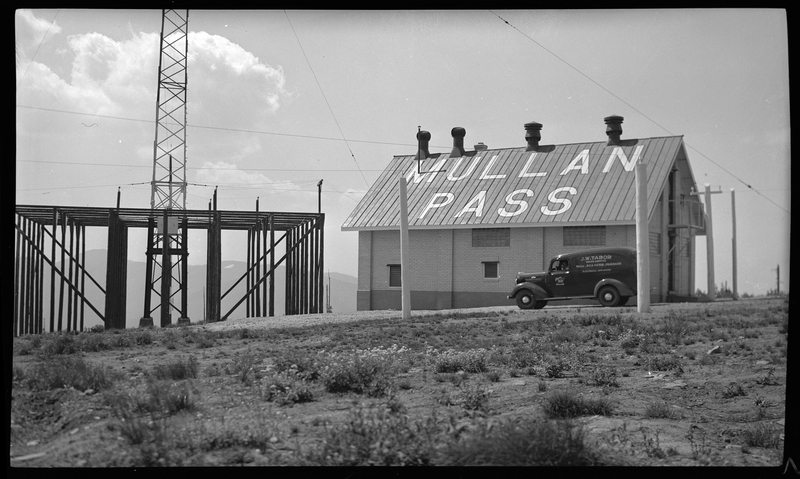Photo of the radio beam station building at Mullan Pass. There is a car parked in front of the building, and the roof of the building has "Mullan Pass" written on it. There is a tall radio tower next to the building.
