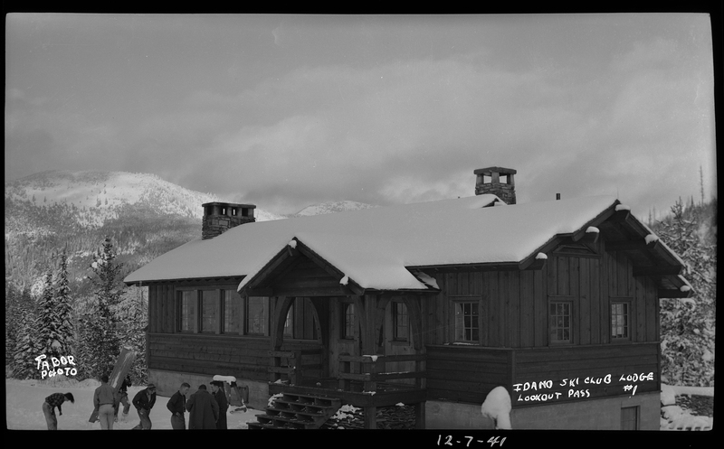 A group of unidentified men are moving something outside the Idaho Ski Club Lodge building in the snow. The ground, roof of the lodge, and the trees in the background are all covered in snow.