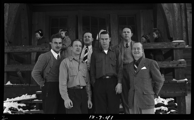 A group of unidentified men stand on the front steps of the Idaho Ski Club Lodge building to pose together for a photo. Four children stand behind them.