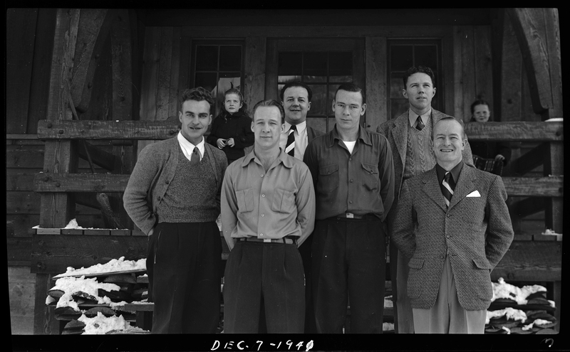 A group of unidentified men stand on the front steps of the Idaho Ski Club Lodge building to pose together for a photo. Two children stand behind them.