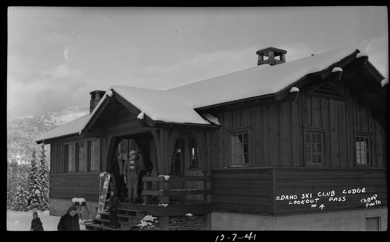 A group of unidentified men and children are standing on the front steps and leaving the Idaho Ski Club Lodge building. There is snow covering the ground and roof of the building.