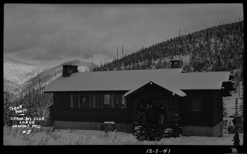 A group of unidentified men are standing on the front steps of the Idaho Ski Club Lodge Building at Lookout Pass. The ground and roof of the building are covered in snow, and trees are visible in the background of the photo.