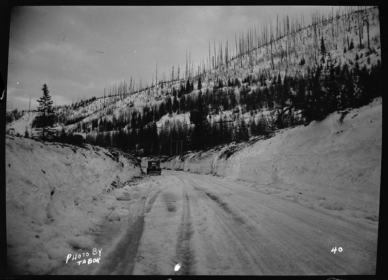 Photo of a car that appears to be parked on the side of the snow covered road near Lookout Pass. There are walls of snow piled up on either side of the road, suggesting regular plowing. Trees can be seen in the background.