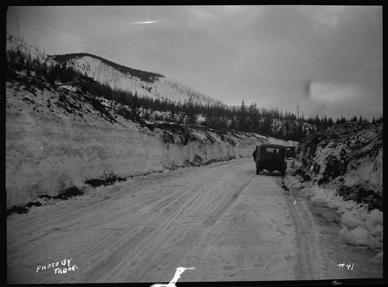 Two cars parked on the side of snow covered road near Lookout Pass. There is an unidentified person mostly concealed by one of the cars. The snow is piled up in walls on both sides of the road, suggesting regular plowing.