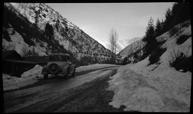 Photo of a car driving down the road (away from the photographer) near Lookout Pass. The road is mostly clear, but there is snow on the sides. There is a building on the other side of the car and trees are seen throughout the landscape.