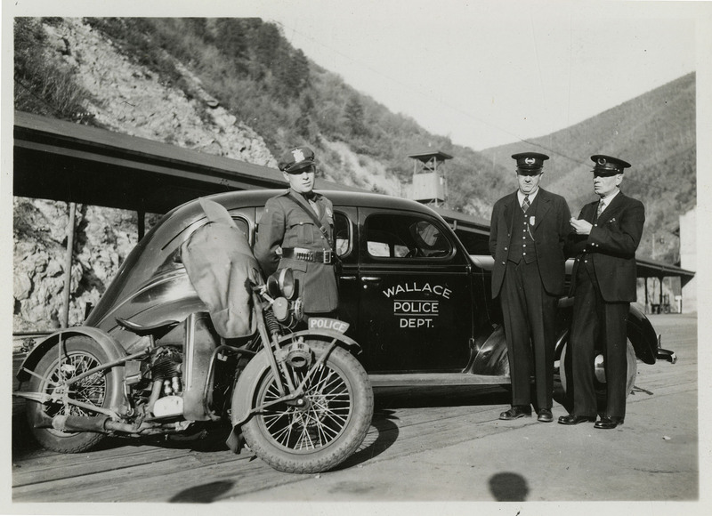 Possibly Wallace police officers in uniform talking to each other while standing near a motorcycle and a Wallace Police Department car outside. Image previously described as: "Tom Barrett, Vic Langley, Charles Pugh. Police photographs." 
