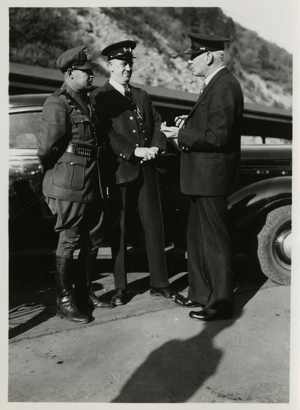 Possibly Wallace police officers in uniform standing near a Wallace Police Department car outside. Image previously described as: "Tom Barrett, Vic Langley, Charles Pugh. Police photographs." 