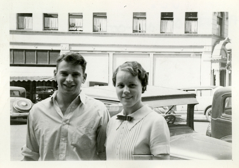 Unidentified man and woman smiling in front of cars parked in the street.