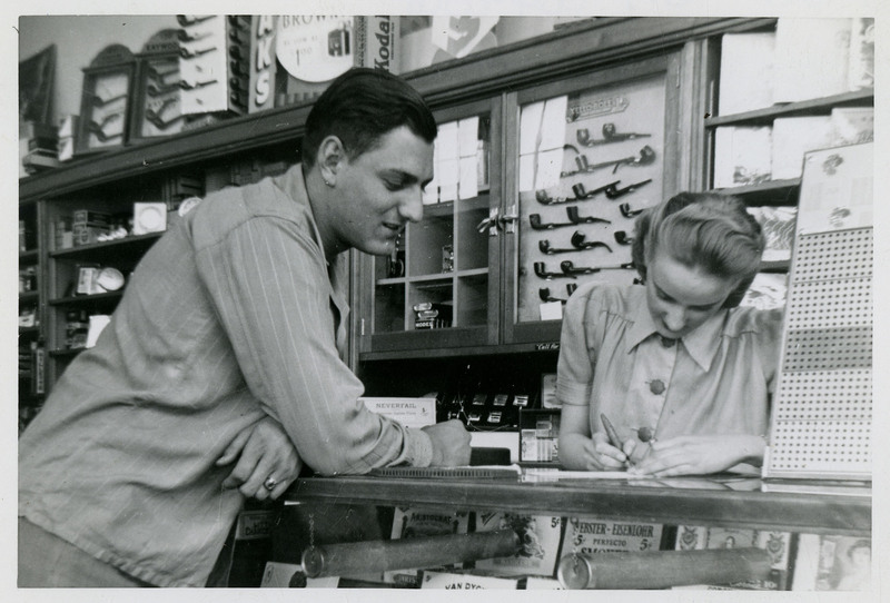 Unidentified man and woman inside a store. The man is watching the woman while she writes behind the counter. There are pipes and other items on display behind the woman and underneath the counter.