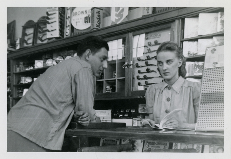 Unidentified man and woman inside a store. The man is watching the woman while she flips through a booklet behind a counter. There are pipes and other items on display behind the woman and underneath the counter.