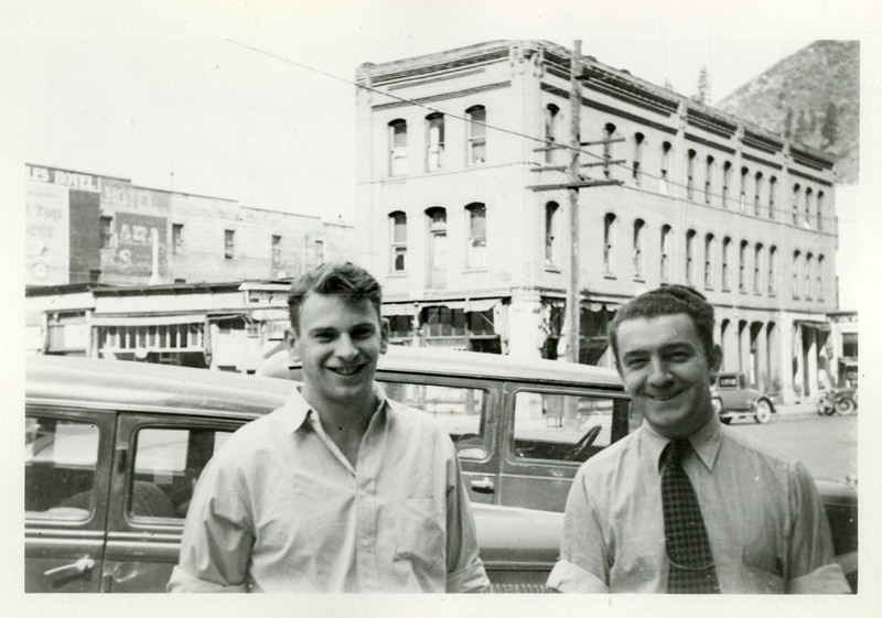 Babe Peila, a photographer for Tabor's, stands on the right of an unidentified man. Both men are smiling at the camera while standing in front of a street.