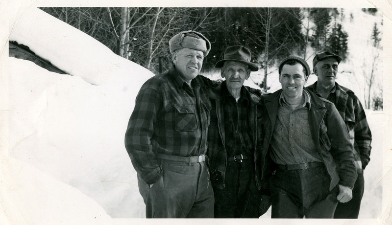 Four unidentified men stand next two each other outside. They are all wearing hats and jackets and there is a thick layer of snow on the ground behind them.
