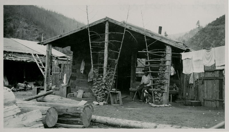 An unidentified man sits on a bench outside a house. A dog is seated next to his leg. There are logs in the foreground and laundry hangs to dry off the side of the house.