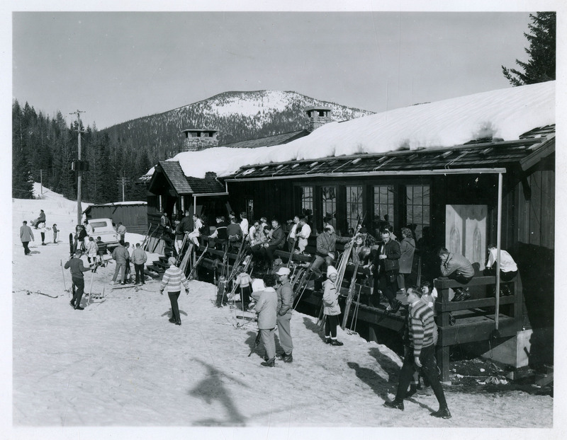 Skiers sit on the railing of a building at the base of Lookout Pass. Skis and ski poles are leaned up against the railing and side of the building. Unidentified people mill about the scene.