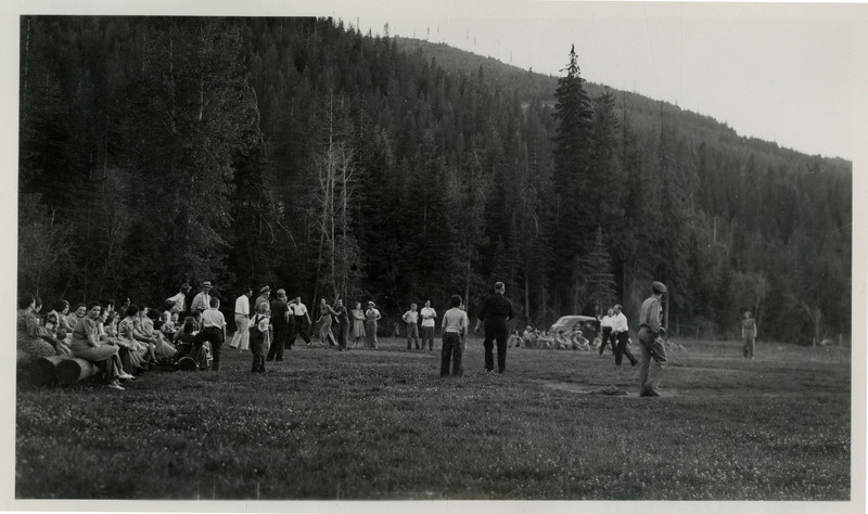A crowd of people mill about a large field. Some are sitting on logs on the sidelines while others appear to play a game. The handwriting on the photo lists several names: [illegible], Lenney, Glen, Mayor, Frank, and Harry.