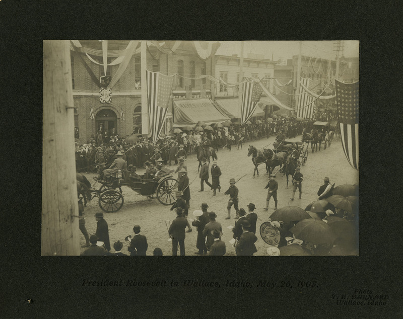 Theodore Roosevelt's procession in Wallace, Idaho turns from 6th street onto Bank street. Armed men separate the crowds from the horse-drawn carriages of the procession. American flags and streamers hang from nearby buildings.