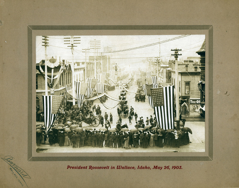 Theodore Roosevelt's procession in Wallace, Idaho. Armed men separate the crowds from the horse-drawn carriages of the procession. American flags and streamers hang from nearby buildings.