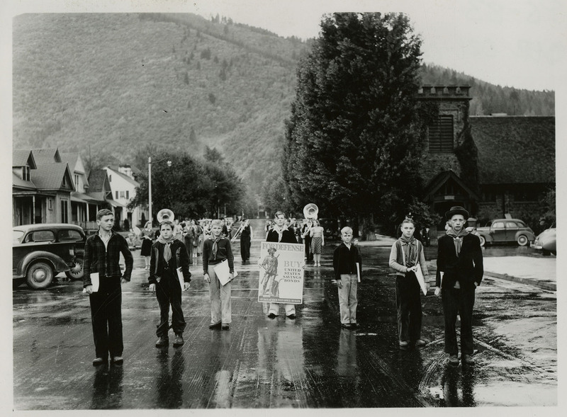 Seven boys stand together as part of the Defense Bond Parade. The boy in the middle is holding a sign that reads "For Defense Buy United States Savings Bonds On Sale At Your Post Office Or Bank."