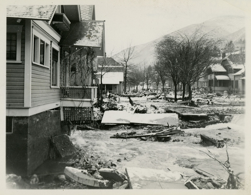 A view of the damage done by a flood in Wallace, Idaho. Several houses and trees are in frame and damaged items are strewn across the ground.