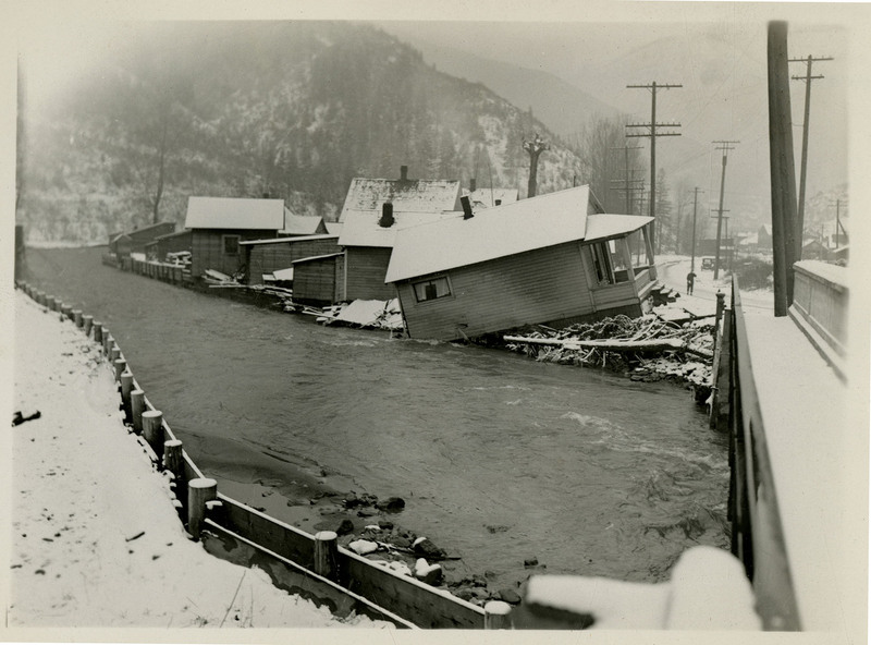 A view of houses near a river after a flood in Wallace, Idaho. There is snow on the ground and one of the houses appears to have a damaged foundation.