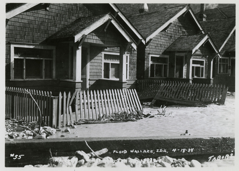 A view of the damage done by a flood in Wallace, Idaho. Two similar houses are in frame, and the fence delineating a front yard has been damaged.
