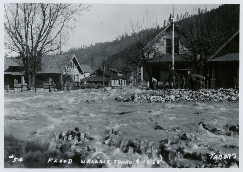 Water rushes past several buildings during a flood in Wallace, Idaho. There is a small group on people on nearby dry land.