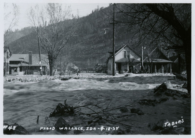 Water rushes past several buildings during a flood in Wallace, Idaho. There are also a few trees in frame and a small group of people on nearby dry land.